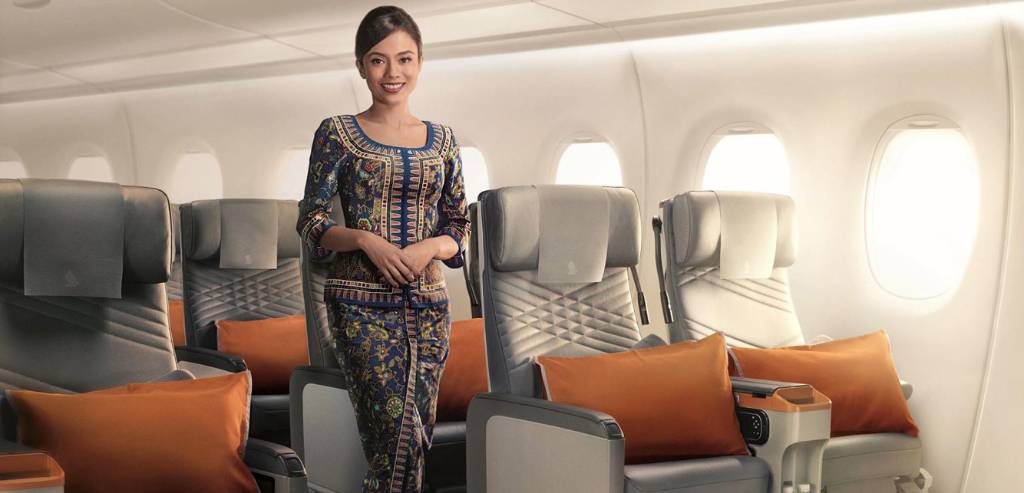 Singapore Airlines premium economy cabin with grey recliners and orange pillows, a cabin crew member standing between the rows